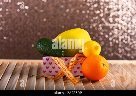 Frutta in scatola con cuori pattern su una superficie di legno. Limone, arancio, melone e di avocado con giallo nastro di misurazione sul beige sfondo bokeh di fondo. Composizione di frutta. Dieta sana e slim concetto di forma Foto Stock
