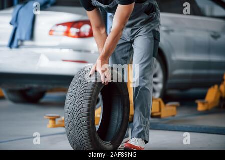 La vettura sul jack dietro. Mechanic detiene un pneumatico presso il garage di riparazione. Sostituzione delle gomme invernali ed estive Foto Stock