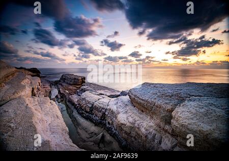 Rocky seashore seascape con oceano ondulata e drammatico e bellissimo tramonto alle grotte marine della zona costiera in Paphos, Cipro Foto Stock