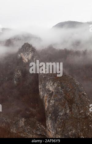 Morbido, misty, spooky vista di forma incredibile scogliera rocciosa, nebbia fitta e rosso, autunno alberi colorati Foto Stock