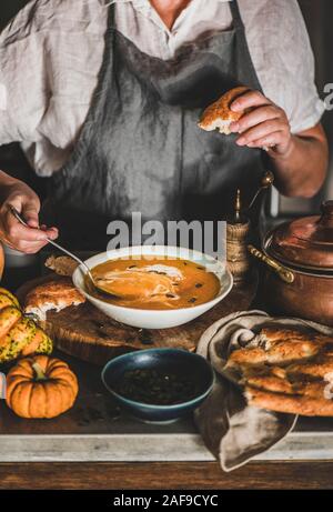 Eldery donna in lino grigio grembiule crema di miscelazione con stagionale autunno Crema di zucca zuppa in cucina moderna con interni. Stagionale autunno comfort food Foto Stock