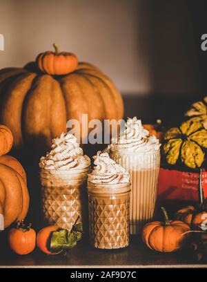 Pumpkin spice latte bevanda caffè la torta con panna montata e cannella in bicchieri alti tra freschi zucche e cachi sul tavolo, parete grigia a bac Foto Stock