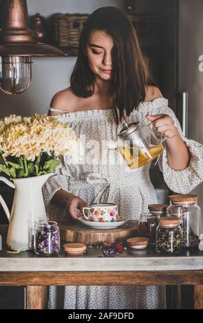 Giovani caucasici sorridente donna con i capelli neri in abito fresco di versamento di infuso di tè verde dal vaso di vetro nella splendida annata tazza di porcellana in cucina Foto Stock