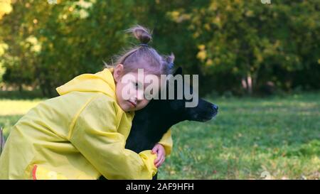 Close-up di una ragazzina in un giallo rainwheel avvolge il suo animale domestico. Si tratta di un cane nero. Foto Stock