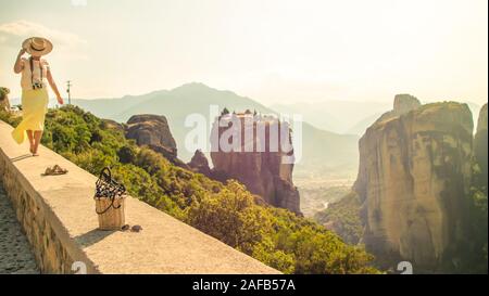 Giovane donna con abito bianco e grandi hat permanente sulla parete di fronte la Grecia meteor montagne, il monastero e il villaggio in background Foto Stock