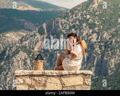 Giovane donna di moda vestire seduta su pietra becnch, fotografare davanti a Delfi paesaggio di montagna in Delphi, Grecia Foto Stock