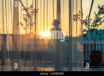 Giovane donna su un balcone guardando il tramonto, vista attraverso le tende trasparenti Foto Stock