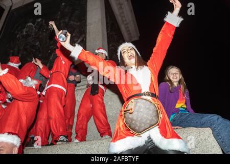Trafalgar Square, Londra, Regno Unito. 14 dicembre 2019. I rivelatori vestiti con costumi di Babbo Natale si riuniscono a Trafalgar Square durante Santacon 2019. La scena notturna cattura una folla vivace che festeggia in abiti festivi, con un partecipante che posa energicamente in primo piano indossando un abito Babbo Natale e una borsa da cintura firmata. Altri si mescolano, ballano e si gusteranno bevande intorno ai gradini del monumento, creando un'atmosfera gioiosa e divertente. L'immagine riflette la giocosa tradizione urbana, l'allegria stagionale e la fusione dell'iconografia natalizia classica con la moderna Street fashion. Penelope Barritt/Alamy Live News Foto Stock