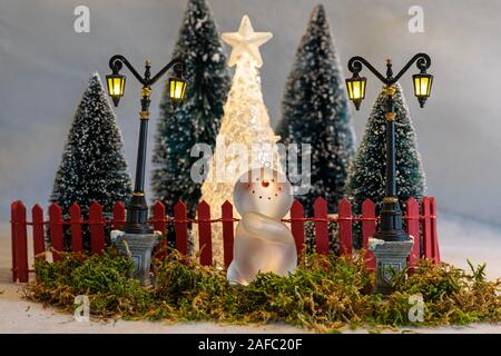 Scheda di Natale, il giocattolo pupazzi di neve (ornamenti) avendo divertimento di fronte ad un giardino recintato con alberi di natale e lanterne Foto Stock