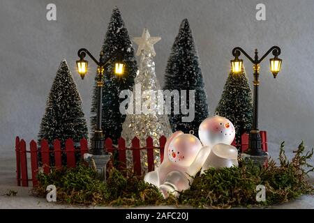 Scheda di Natale, il giocattolo pupazzi di neve (ornamenti) avendo divertimento di fronte ad un giardino recintato con alberi di natale e lanterne Foto Stock