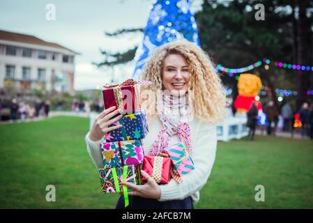 Sorridendo felice bionda donna ricci con torre di colorati confezioni regalo in mani, godersi il tempo all'aperto nel parco decorata con alleggerita albero di Natale o Foto Stock