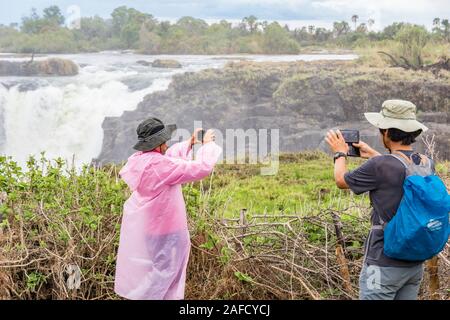 Cascate Victoria, Zimbabwe. Due escursionisti scattano foto delle cascate. L'isola di Livingstone si può vedere sullo sfondo. Foto Stock