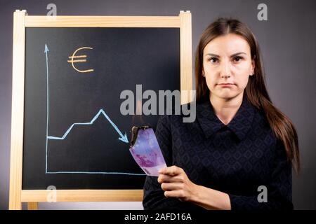 Il grafico della caduta dell' euro è disegnato su una lavagna nera, un triste donna è in piedi accanto a lei e che è in possesso di una masterizzazione nota nella quantità di Foto Stock