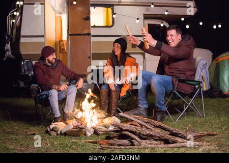 Gruppo di amici in fase di riscaldamento intorno al fuoco di campo e ridere insieme. Retrò camper. Foto Stock