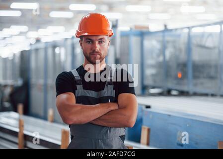 Gli stand con le braccia incrociate. Ritratto del maschio lavoratore industriale in ambienti interni in fabbrica. Giovane tecnico con orange elmetto Foto Stock