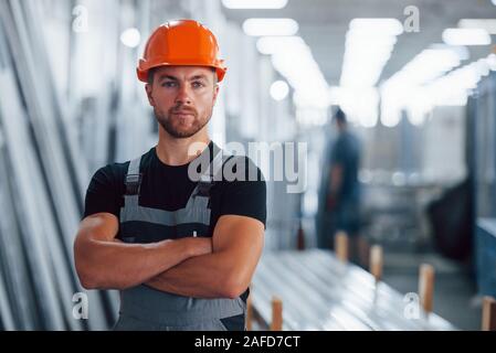 Gli stand con le braccia incrociate. Ritratto del maschio lavoratore industriale in ambienti interni in fabbrica. Giovane tecnico con orange elmetto Foto Stock