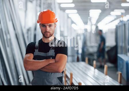 Gli stand con le braccia incrociate. Ritratto del maschio lavoratore industriale in ambienti interni in fabbrica. Giovane tecnico con orange elmetto Foto Stock