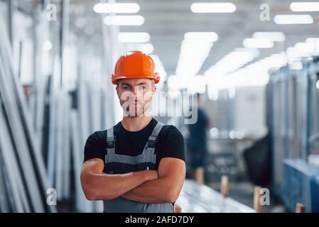 Gli stand con le braccia incrociate. Ritratto del maschio lavoratore industriale in ambienti interni in fabbrica. Giovane tecnico con orange elmetto Foto Stock