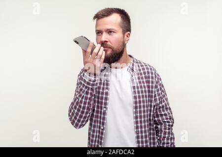 Assistente virtuale. Ritratto di grave uomo barbuto in casuale plaid shirt parlando al telefono mobile utilizzando applicazioni vocali, un messaggio audio o registratore. i Foto Stock