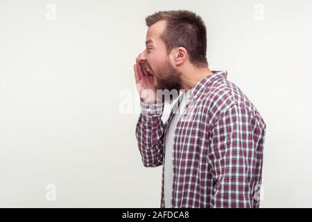 Vista laterale di arrabbiato barbuto Uomo in camicia casual gridare a gran voce il braccio di contenimento in prossimità della bocca, gridando con espressione aggressiva, luogo vuoto per il testo pubblicitario, co Foto Stock