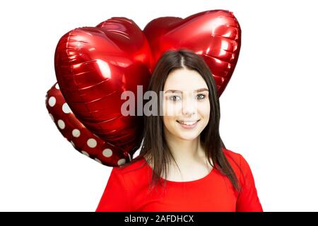 Una bruna ragazza in un abito rosso detiene oltre la sua testa tre rosso a forma di cuore palloncini per il giorno di San Valentino Foto Stock