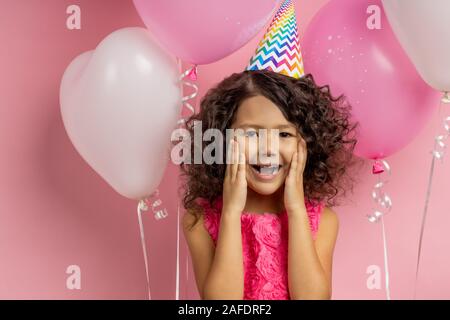 Che sorpresa. Carino contentissimo caucasian bambina abito, party hat, che si erge tra i palloncini con bocca aperta, mantenendo le palme sulle verifiche, ricezione Foto Stock