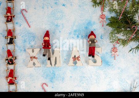 Little dwarf bambole sono seduti sulle scale di Natale. Le decorazioni di Natale in legno per lettere XMAS su un blu e sfondo bianco Foto Stock