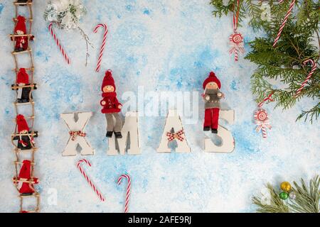 Little dwarf bambole sono seduti sulle scale di Natale. Le decorazioni di Natale in legno per lettere XMAS su un blu e sfondo bianco Foto Stock