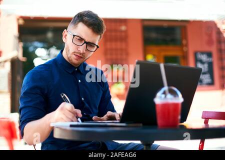 Libero professionista man mano che scrive sul notebook di carta vicino al suo laptop in outdoor cafe. Giovane imprenditore in bicchieri prendendo appunti nel Blocco note con penna elegante Foto Stock