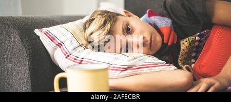 Ragazzo malato giacente con coperta di lana, bottiglie di acqua calda e di una tazza alta. Triste teen con l'influenza si appoggia a casa in una fredda giornata invernale. Bambino con infectio stagionali Foto Stock