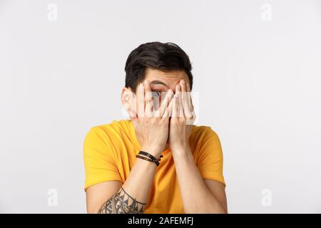 Spaventata ma curioso carino uomo asiatico con corti capelli scuri, tatuaggi e nasconde il viso, chiudere gli occhi con le mani e spiata attraverso le dita in corrispondenza importante intenso Foto Stock