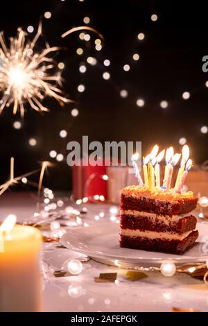 Pezzo di torta con molte candele accese sulla piastra circondato da luci scintillanti Foto Stock