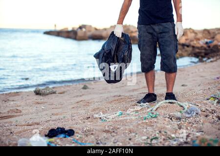 Uomo in maglietta scura e pantaloncini con guanti bianchi e big black package raccogliere rifiuti sulla spiaggia. La tutela ambientale e il pianeta inquinamento co Foto Stock