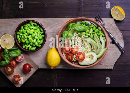 Una fresca e sana insalata con zucchine zoodles tagliatelle, baby pomodori, avocado e fagioli edamame. L'insalata è in una ciotola di legno al buio su un tavolo di bambù o Foto Stock