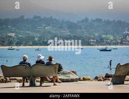 Persone sedersi sulla coda di balena panca si affaccia la chiesa di Santa Barbara del porto con SUPB, barche a vela e le prime colline nella nebbia, a Santa Barbara, CA, Stati Uniti d'America Foto Stock