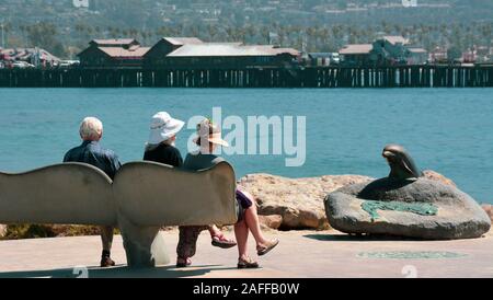 Persone sedersi sul racconto di balena panche si affaccia la chiesa di Santa Barbara Porto di bronzo con una scultura di delfini e Poppe Wharf, Santa Barbara, CA, Stati Uniti d'America Foto Stock