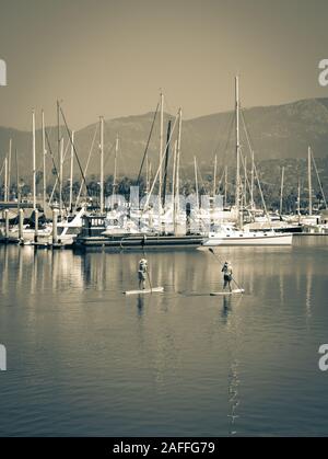 Due donne maneuverer loro stand up paddle boards attraverso un canale vicino ormeggiata barche a vela e yacht in marina nella Santa Barbara, Porto Santa Foto Stock