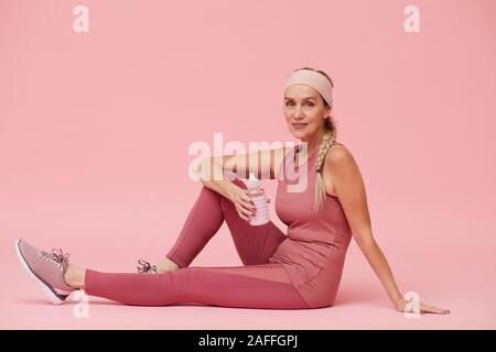 A piena lunghezza Ritratto di montare donna matura tenendo la bottiglia di acqua mentre è seduto sul pavimento contro di rosa pastello, sfondo spazio copia Foto Stock