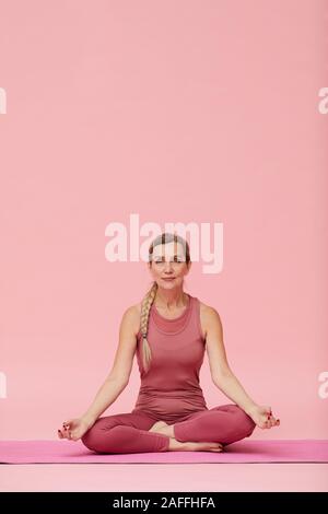A piena lunghezza Ritratto di montare donna matura fare yoga contro di rosa pastello, sfondo posizione del loto Foto Stock