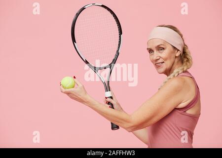 Vita ritratto di active donna matura azienda racchetta da tennis mentre in piedi contro di rosa pastello, sfondo spazio copia Foto Stock