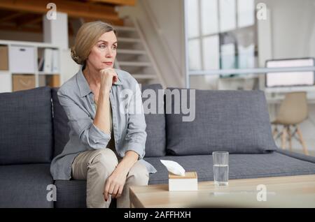Ritratto di eleganti donna matura seduti sul divano di casa e guardando lontano pensively, triste o depressa con tessuti su tavola, spazio copia Foto Stock