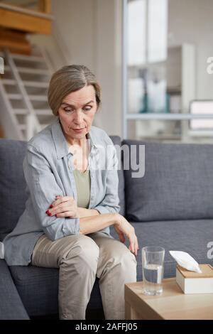 Ritratto di triste donna matura seduti sul divano di casa e guardando verso il basso con la faccia triste espressione, tessuti e acqua sul tavolo, spazio di copia Foto Stock