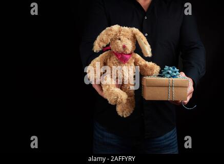 Adulto Uomo sorge su uno sfondo scuro che indossa una camicia nera e tiene in mano un marrone scatola di cartone con un dono e un simpatico coniglio marrone toy, felice Foto Stock