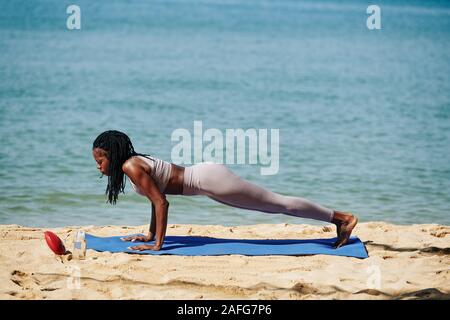 Concentrato di giovane donna nero facendo push-up sul materassino yoga sulla spiaggia di mattina Foto Stock