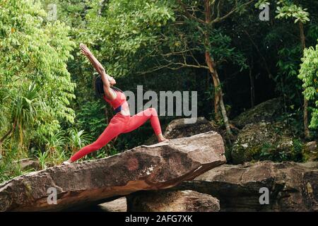 Slim giovane donna in piedi sulla roccia nella foresta tropicale e facendo warrior pongono Foto Stock
