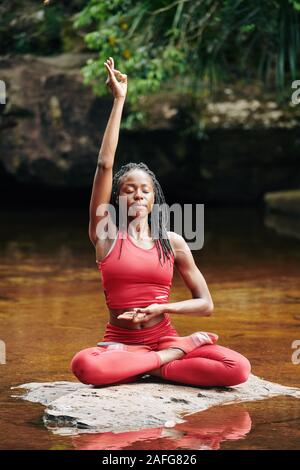 Concentrato di giovane donna nero facendo gyan mudra quando si è seduti sulla roccia nella posizione del loto Foto Stock