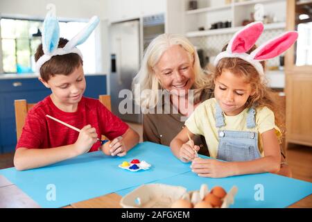 La nonna con i nipoti indossando orecchie di coniglio decorare le uova di Pasqua in casa insieme Foto Stock