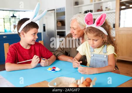 La nonna con i nipoti indossando orecchie di coniglio decorare le uova di Pasqua in casa insieme Foto Stock