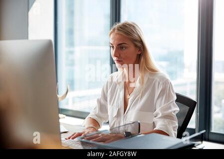 Bianco giovane donna bionda seduti ad una scrivania che lavora a un computer in un ufficio creativo, close up Foto Stock