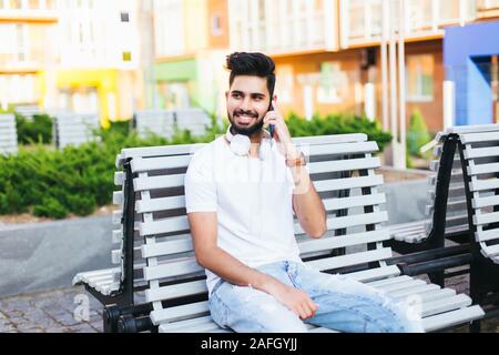 Giovani indiani uomo seduto su un banco di lavoro, parla al telefono, guardando la fotocamera. Uomo bello in bicchieri di indossare in bianco maglietta elegante. Lavoratore it sorridente, pos Foto Stock